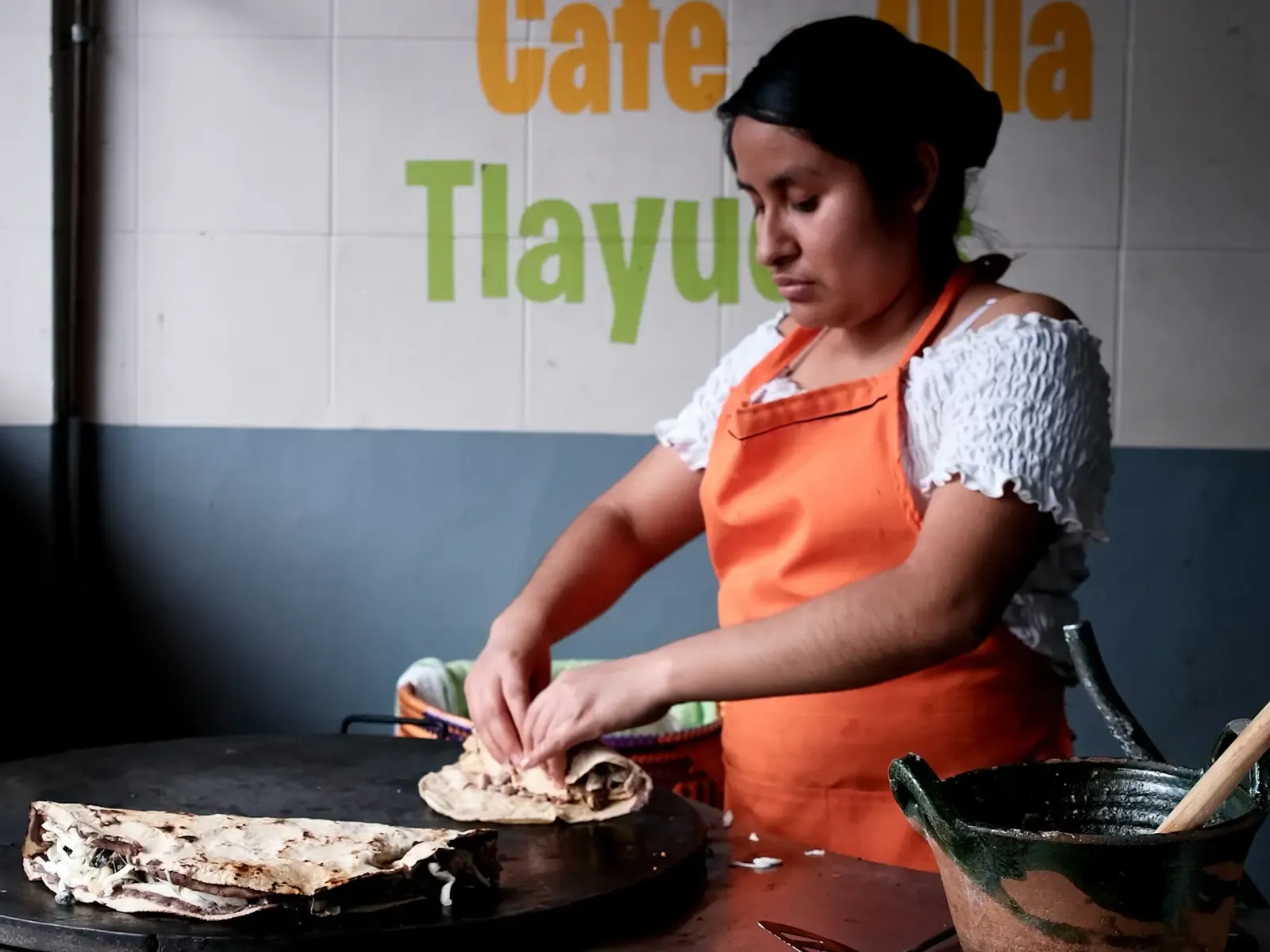 Cocinera con delantal anaranjado doblando una tortilla sobre un comal grande