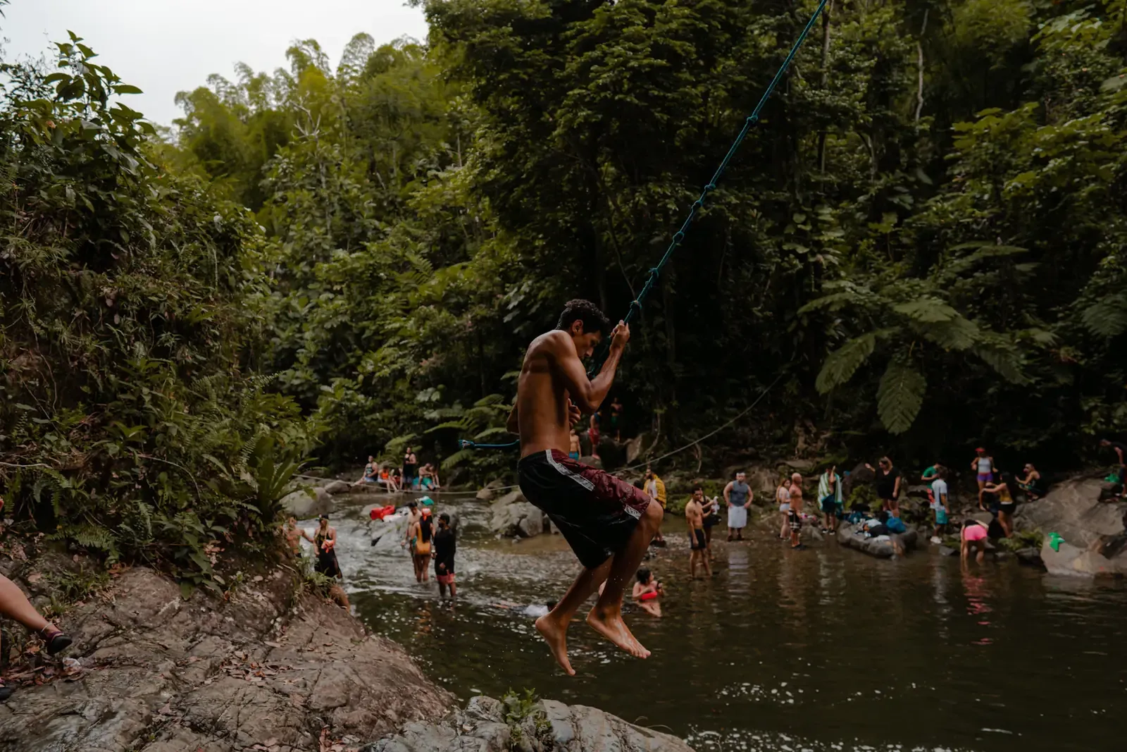 Persona lanzándose desde un columpio de cuerda a un río en medio del bosque, con más gente bañándose